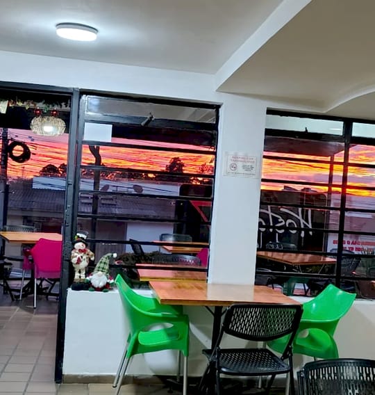 Mesas de madera y sillas verdes junto a los ventanales; afuera, el cielo de Manizales encendido en naranja y rosa al atardecer.