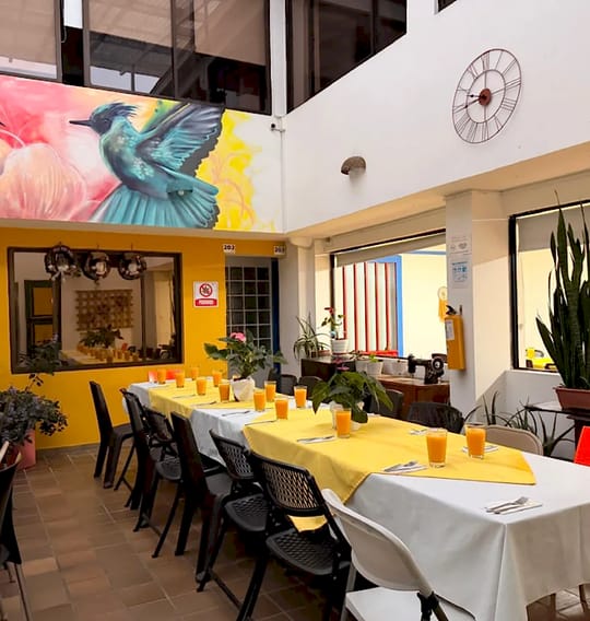 El comedor desde otro ángulo: colibrí turquesa pintado sobre la pared amarilla, reloj de pared, puertas de las habitaciones 202 y 203 y mesas preparadas.