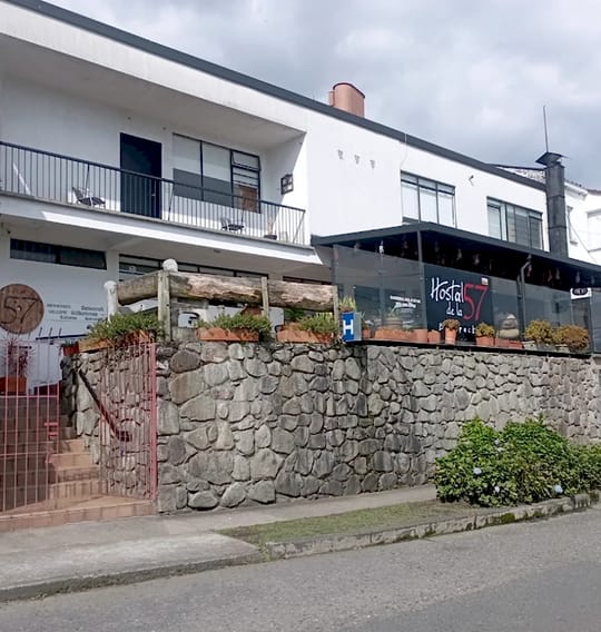 Fachada blanca de dos pisos con balcón, muro de piedra con jardineras, escalera con reja roja y el letrero negro «Hostal de la 57».