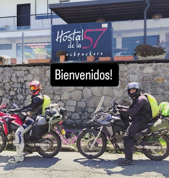 Dos motoviajeros con casco y motos cargadas frente al letrero «Hostal de la 57 backpackers» y el muro de piedra; sobre la foto, el texto «Bienvenidos!».