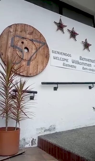 Muro blanco de la entrada con un carrete de madera pintado con el número 57 y un letrero de bienvenida en seis idiomas.