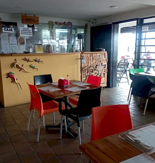 Interior del restaurante: mesa con sillas naranja y negras, mostrador de caja amarillo con lagartijas decorativas y puerta abierta hacia la terraza.