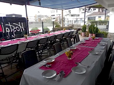 Mesa larga en la terraza con manteles blancos, caminos fucsia, platos y flores, lista para una celebración.